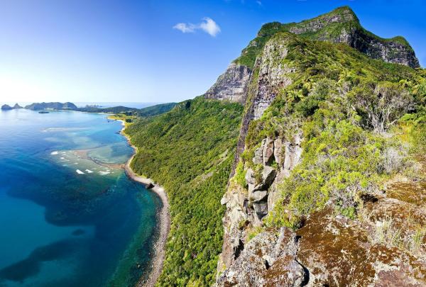 A breathtaking panoramic view from the rocky summit of Mount Gower at Capella Lodge on Lord Howe Island, looking down at the lush green slopes, turquoise coral lagoon, and the distant, winding coastline under a clear blue sky. © Capella Lodge