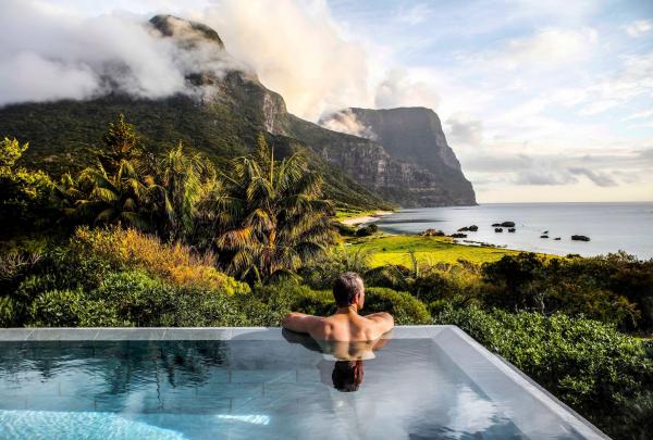 A man relaxes in a crystal-clear infinity pool at Capella Lodge on Lord Howe Island, gazing out at the lush tropical greenery and the dramatic, cloud-shrouded peaks of Mount Gower and Mount Lidgbird by the calm sea. © Bailie Lodges