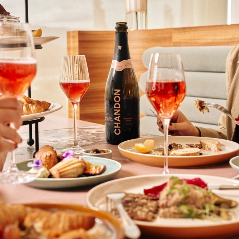 An elegant high tea spread at Chandon in Coldstream, Victoria, features a bottle of sparkling rosé alongside tiered platters of pastries and savoury bites, accompanied by filled wine glasses in a bright, modern dining room. © Tourism Australia