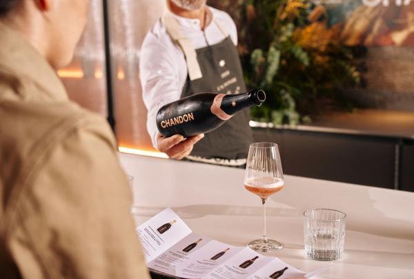 A server at Chandon in Coldstream, Victoria, pours a glass of chilled sparkling rosé for a guest at a sleek white tasting bar, with a detailed wine menu and elegant floral arrangements completing the refined Yarra Valley experience. © Tourism Australia