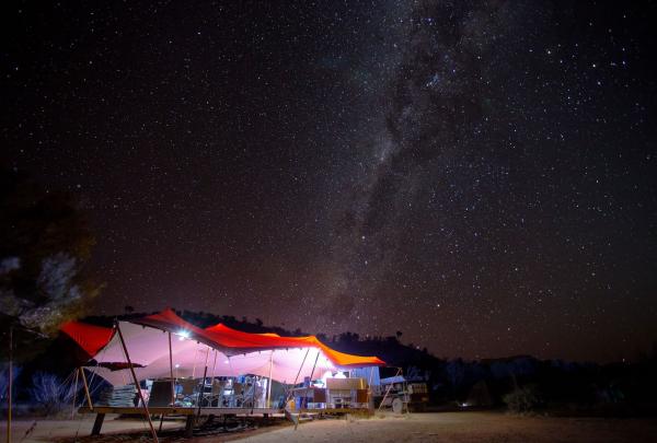 Under a vast, star-filled outback sky, the Milky Way stretches brilliantly over a glowingly lit, open-air safari tent at the Classic Larapinta Trek in Comfort in the Northern Territory, creating a magical nocturnal scene. © World Expeditions/Great Walks of Australia