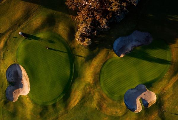 An overhead view of the Commonwealth Golf Club in Melbourne, Victoria, shows two perfectly manicured circular greens punctuated by deep, white-sand bunkers, with long shadows cast across the grass by the warm, golden afternoon sun. © Great Golf Courses of Australia