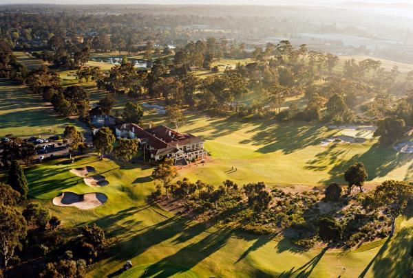 An aerial view of the Commonwealth Golf Club in Melbourne, Victoria, showcases lush green fairways and sculptural sand bunkers weaving through a dense landscape of native trees, all bathed in the warm, golden light of a low sun. © Great Golf Courses of Australia