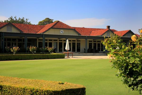 The historic clubhouse at Concord Golf Club in Sydney, New South Wales, features a distinctive red-tiled roof and a broad, shaded veranda overlooking a perfectly manicured green lawn and vibrant, blooming rose bushes. © Concord Golf Club