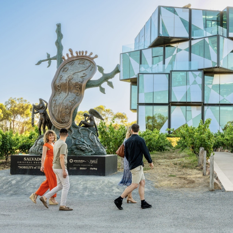 Four visitors walk past a large, surrealist bronze sculpture of a melting clock toward the geometric, glass-paneled d'Arenberg Cube in McLaren Vale, South Australia, which stands strikingly amidst the surrounding green vineyards. © Tourism Australia