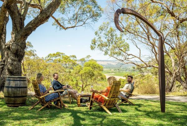 Four people sit in rustic wooden chairs under the shade of large, leafy trees at d'Arenberg in McLaren Vale, South Australia, enjoying glasses of wine with a view of the rolling green valley and a tall, weathered metal sculpture in the foreground. © Tourism Australia