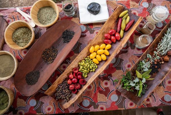 An overhead view of a bush tucker display at Dale Tilbrook Experiences in Perth shows traditional wooden coolamons filled with native Australian ingredients. The spread includes colorful finger limes, desert limes, quandongs, and various seeds and spices arranged on an Aboriginal-patterned cloth. © Tourism Australia