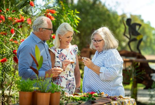 Dale Tilbrook and two guests sample native ingredients among red bottlebrush flowers and lush greenery in a sunny garden at Dale Tilbrook Experiences in Perth, Western Australia. © Tourism Australia