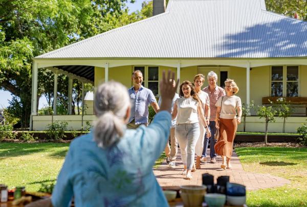 In a sun-drenched garden at Dale Tilbrook Experiences in Perth, a group of five visitors walks along a path toward a welcoming host. A grand, white weatherboard house with a sprawling veranda and a silvery corrugated metal roof is seen in the background. © Tourism Australia © Tourism Australia