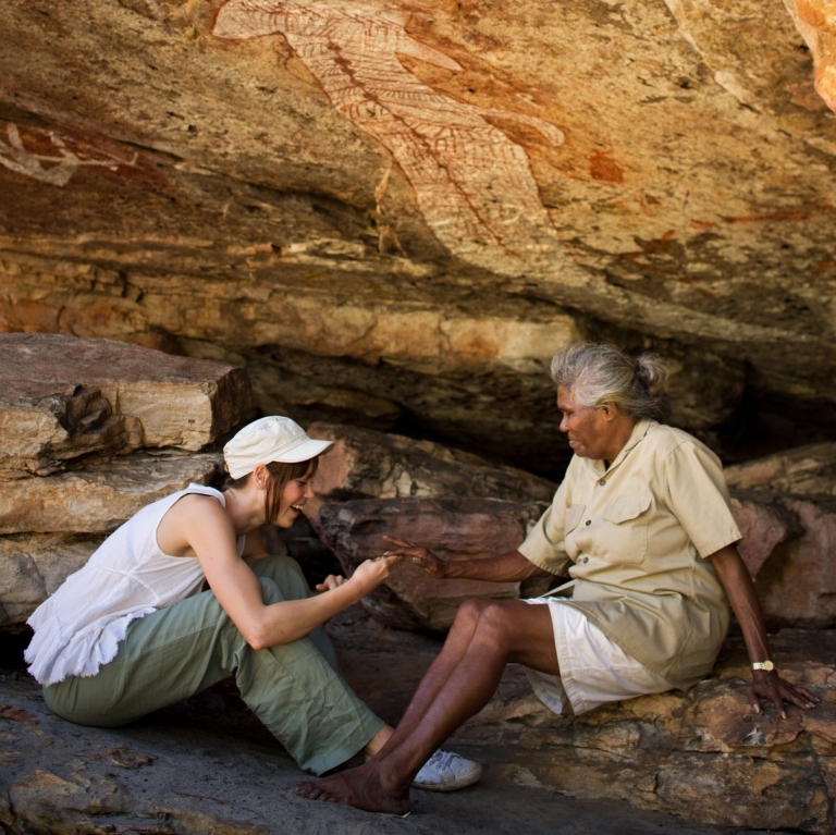 At Davidsons Arnhemland Safaris, an Aboriginal guide and a guest sit beneath a rock overhang, sharing stories near a large, ancient ochre painting of a barramundi fish on the ceiling in West Arnhem Land, Northern Territory. © Tourism Australia