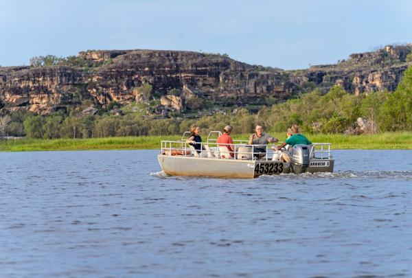 A group of guests enjoys a boat cruise across a calm billabong, framed by rugged sandstone escarpments and lush green wetlands in the distance, as part of Davidsons Arnhemland Safaris in West Arnhem Land, Northern Territory. © Tourism Australia