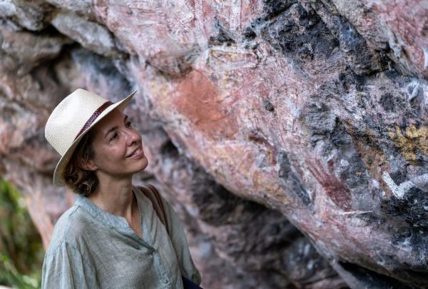 A smiling woman in a sun hat gazes at ancient, colourful Indigenous rock art painted on a textured stone overhang during a guided outdoor adventure with Davidsons Arnhemland Safaris in West Arnhem Land, Northern Territory. © Tourism Australia