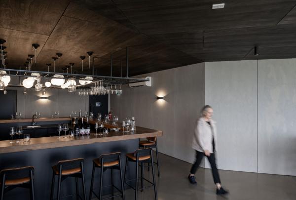 Inside the modern Devil’s Corner cellar door on the East Coast of Tasmania, a woman walks past a stylish black bar featuring a timber countertop, leather-seated stools, and a unique overhead rack filled with wine glasses. © Tourism Australia