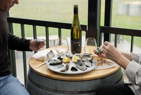 Sitting on a rainy deck at Devil’s Corner on the East Coast of Tasmania, two people share a platter of fresh oysters and a bottle of white wine on a rustic barrel table, overlooking the misty, green vineyard. © Tourism Australia