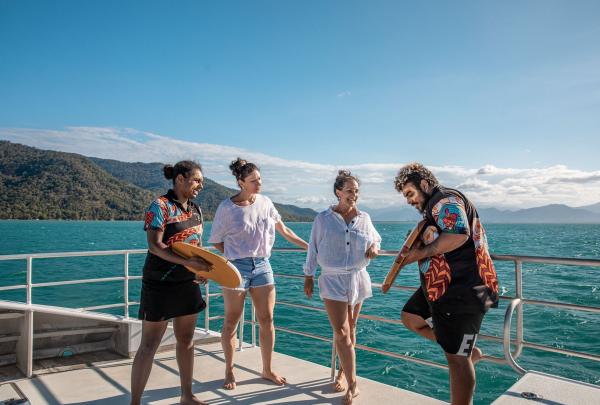 Image Alt Text Custom Gem Image Alt Text said On the deck of a Dreamtime Dive & Snorkel boat on the Great Barrier Reef in Queensland, Indigenous guides in vibrant uniform shirts share cultural stories and traditional instruments with two interested female passengers. © Tourism and Events Queensland