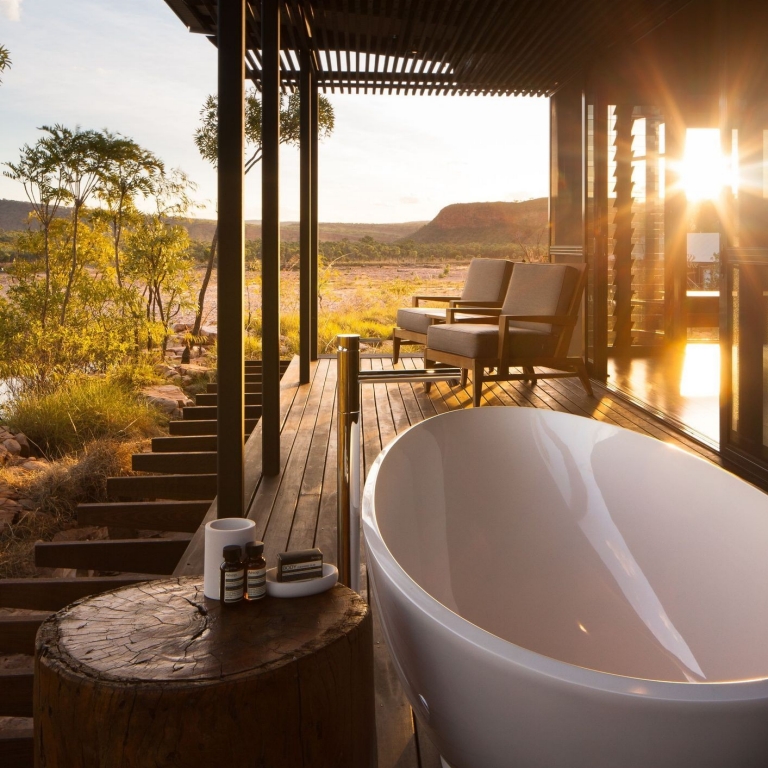 A sleek, white freestanding bathtub sits on a private timber deck at El Questro Homestead in the Kimberley, Western Australia. The golden setting sun glows through the slats, overlooking the vast, rugged outback landscape. © Delware North Companies
