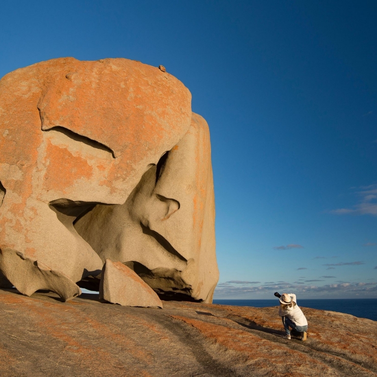 A photographer crouches to capture the iconic Remarkable Rocks with Exceptional Kangaroo Island, Kangaroo Island, South Australia. The massive, weathered granite boulders are covered in orange lichen, set against a deep blue sky and ocean. © Exceptional Kangaroo Island/Tourism Australia