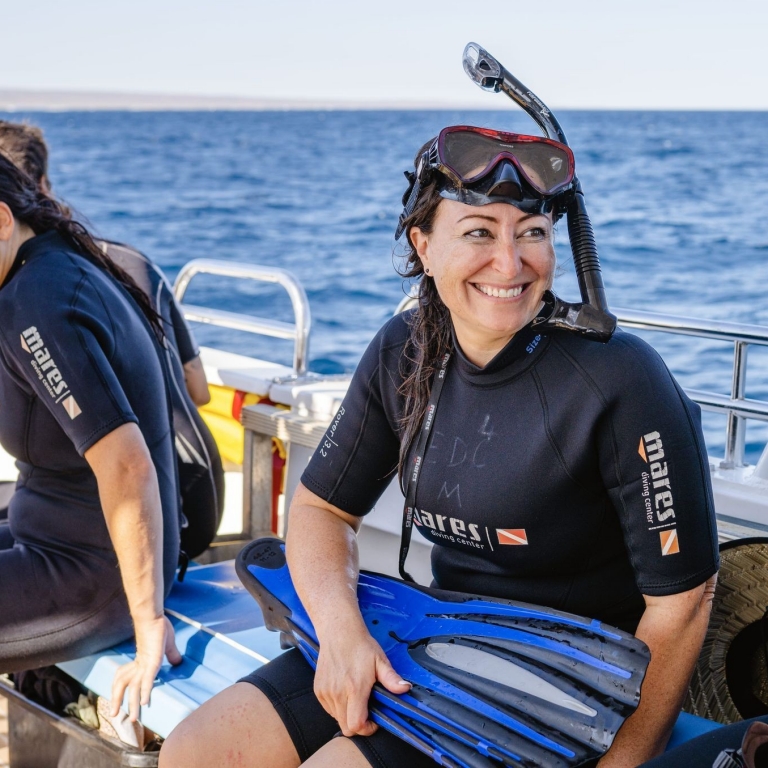 A smiling man and woman in wetsuits sit on a boat with Exmouth Dive and Whalesharks Ningaloo, Ningaloo Reef, Western Australia. They are preparing for a swim, holding snorkelling fins against a backdrop of sparkling blue ocean. © Tourism Australia