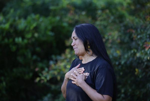 An Aboriginal guide from Explore Byron Bay in New South Wales stands with her hands over her heart, a gesture of connection. She wears traditional-style earrings and a black tour shirt, set against a soft-focus green forest. © Tourism Australia