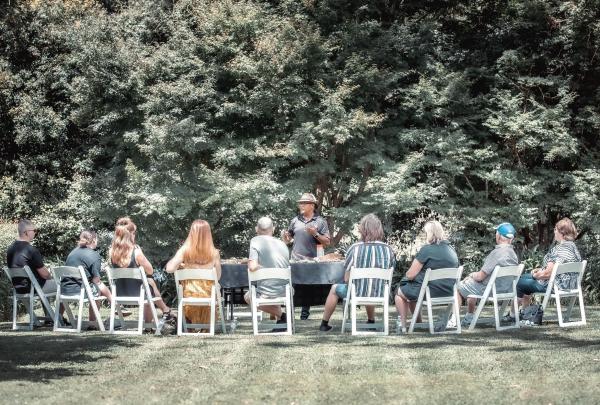 Image Alt Text Custom Gem Image Alt Text said An Aboriginal guide leads an outdoor bush tucker talk for a group of seated guests at Firescreek Botanical Winery on the Central Coast, New South Wales. The group sits in white chairs on a lush green lawn, shaded by dense trees. © Tourism Australia