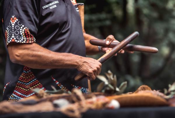 An Aboriginal guide demonstrates the use of traditional wooden clapsticks at Firescreek Botanical Winery, Central Coast, New South Wales. He wears a black polo shirt featuring intricate Indigenous dot art on the side and sleeve. © Tourism Australia