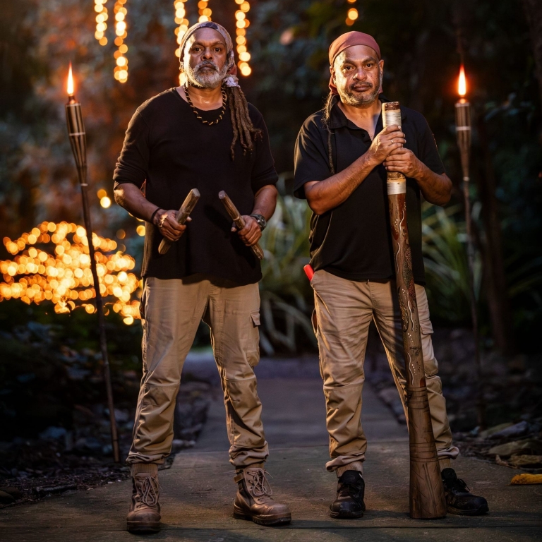 Two Aboriginal guides stand on a path at Flames of the Forest in Cairns, Queensland. One holds traditional clapsticks and the other a didgeridoo, flanked by flaming torches with golden fairy lights twinkling in the rainforest. © Tourism Australia