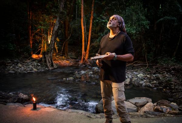 An Aboriginal guide stands by a rocky stream during the Flames of the Forest experience in Cairns, Queensland. Clad in a black shirt and bandana, he looks upward, holding traditional clapsticks amidst the floodlit rainforest. © Tourism Australia