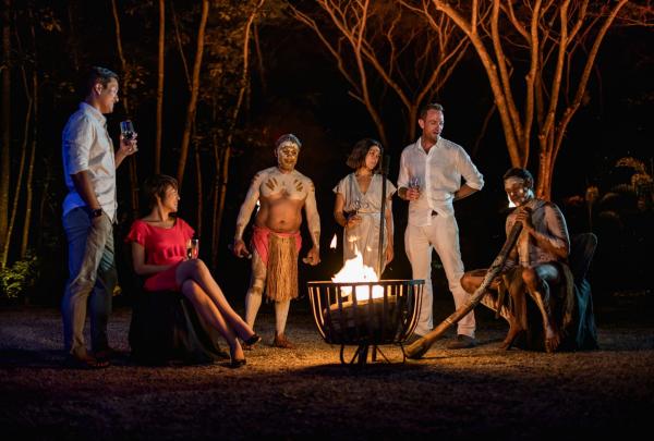 A group of guests gathers around a fire pit during an evening Flames of the Forest experience in Cairns, Queensland. Aboriginal performers in traditional body paint share stories and play the didgeridoo in the rainforest. © Tourism Australia