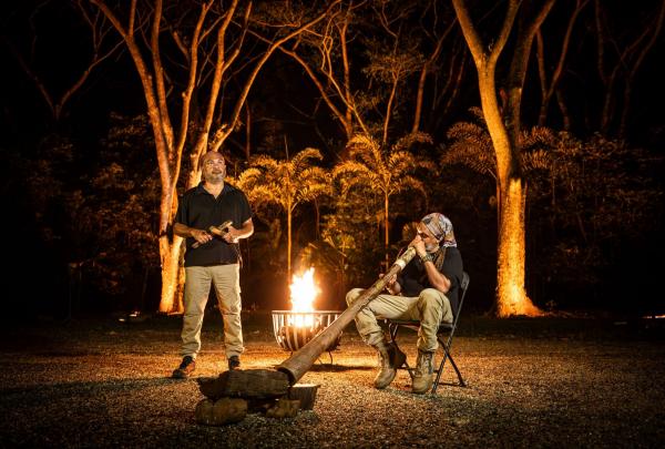 Two Aboriginal guides perform by a crackling fire during a nighttime Flames of the Forest experience in Cairns, Queensland. One man plays a didgeridoo while the other stands nearby, surrounded by towering, floodlit rainforest trees. © Tourism Australia