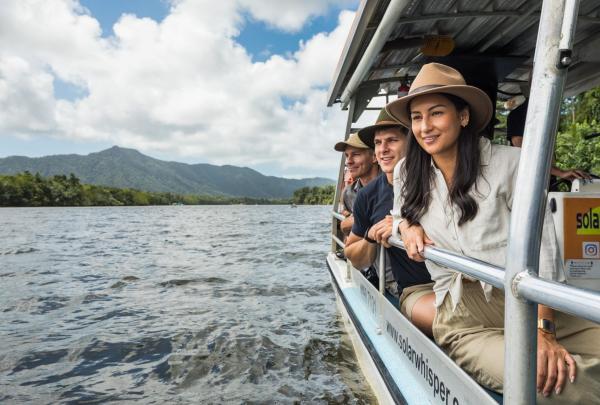 Guests sit on a boat during an FNQ Nature Tours cruise on the Daintree River in Queensland, looking out over the calm water toward lush, green rainforest mountains. © Tourism Australia