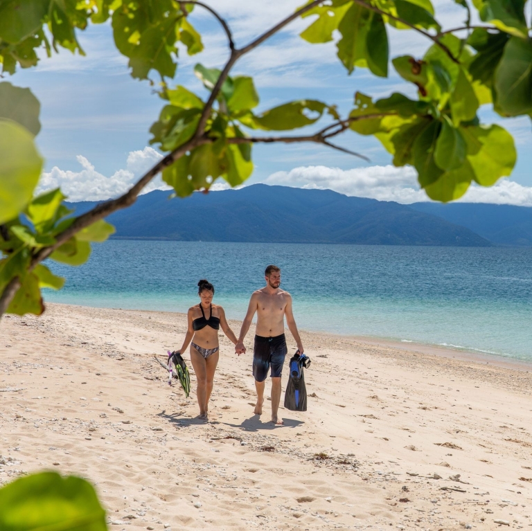 A couple walks hand-in-hand along the pristine sandy shore of the Frankland Islands, Queensland, carrying snorkelling gear. © Tourism Australia