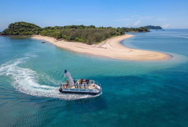 An aerial view shows a white Frankland Islands Reef Cruises boat cruising through turquoise water past a pristine sandy beach on the Frankland Islands, Queensland. The island is covered in lush green trees under a clear blue sky. © Frankland Islands Reef Cruises