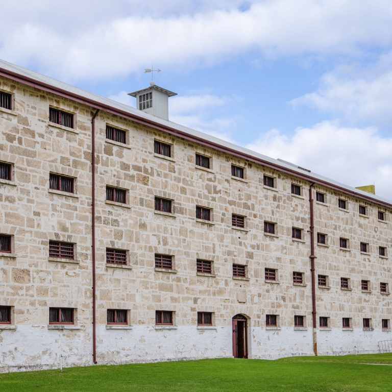 The imposing limestone façade of Fremantle Prison in Perth, Western Australia, features rows of small, barred cell windows under a cloudy sky. This historic site offers a glimpse into the convict history of Western Australia. © Tourism Australia