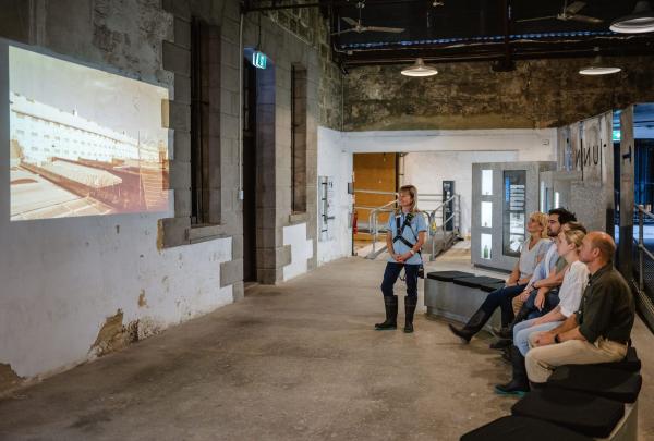 A group of visitors sits in a rustic limestone room at Fremantle Prison, Perth, watching a historical projection on a weathered wall. A guide in a blue uniform stands nearby during this engaging Western Australia heritage tour. © Tourism Australia