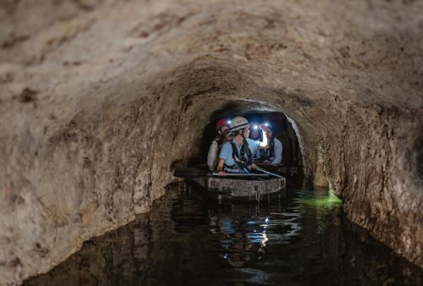 In a small boat, a group of visitors wearing helmets and headlamps navigates a narrow, water-filled underground passage at Fremantle Prison, Perth. This Tunnels tour highlights the limestone walls of the Western Australia site. © Tourism Australia