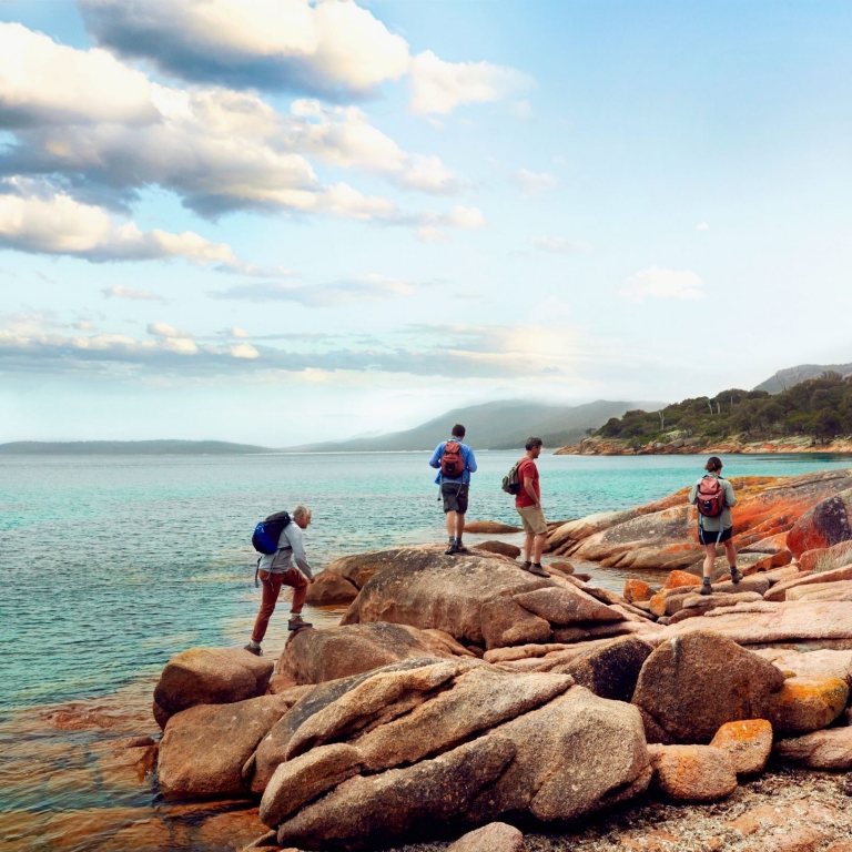 Four hikers with backpacks explore the rugged coastline during a Freycinet Experience Walk in Tasmania. They navigate large, orange-toned granite boulders at the water's edge, overlooking the calm turquoise ocean under a soft, clouded sky. © Tourism Australia