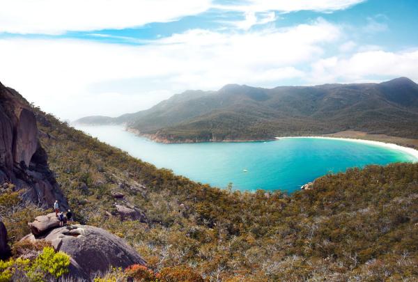 From a rocky outcrop, hikers overlook the bright turquoise waters of Wineglass Bay during the Freycinet Experience Walk in Tasmania. The white sand beach is framed by dense green bushland and the rolling granite peaks of the Hazards. © Tourism Australia