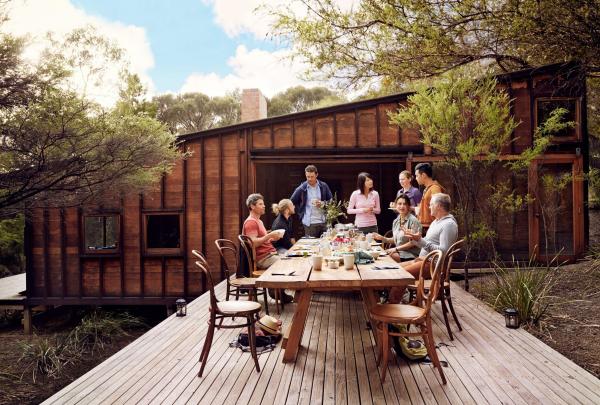 A group of guests enjoys an outdoor meal at a long wooden table on a spacious deck during the Freycinet Experience Walk in Tasmania. The rustic timber lodge is nestled among native trees, offering a serene bush setting for hikers. © Tourism Australia