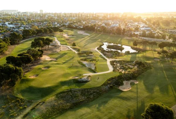 An aerial view captures the strategic layout of Glenelg Golf Club in South Australia, highlighting its manicured green fairways, sandy bunkers, and serene wetlands. The course is nestled within a residential suburb near the coast under the warm glow of a setting sun. © Glenelg Golf Club