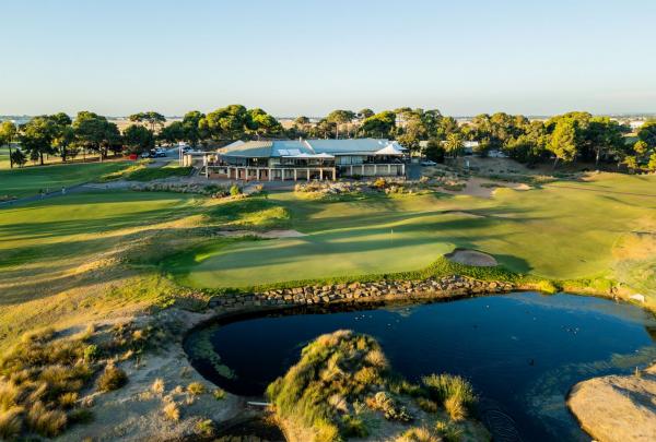 An aerial view of the Glenelg Golf Club in South Australia shows the modern clubhouse overlooking a lush green course. A calm water hazard sits in the foreground, reflecting the golden afternoon light across the fairways and surrounding trees. © Glenelg Golf Club