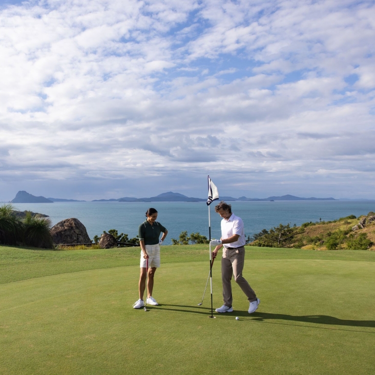 Two golfers stand on a bright green at Hamilton Island Golf Club, Queensland. One player attends to the flagstick while looking out over the expansive blue ocean and the silhouettes of distant islands in the Whitsundays. © Tourism Australia