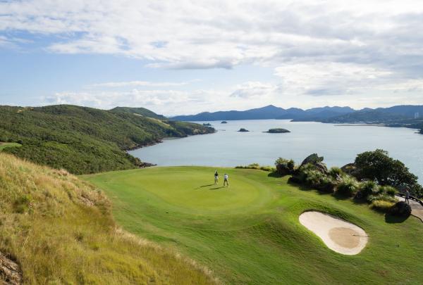 Two golfers walk across a pristine green at Hamilton Island Golf Club, Queensland. The course is perched on a cliff edge, offering breathtaking views over the turquoise waters and tropical islands of the Whitsundays under a bright sky. © Tourism Australia
