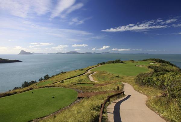 A winding concrete path leads through grassy hills toward a bright green fairway at Hamilton Island Golf Club, Queensland. The course is surrounded by the vast blue ocean and distant tropical islands of the Whitsundays under a clear sky. © Hamilton Island Golf Club