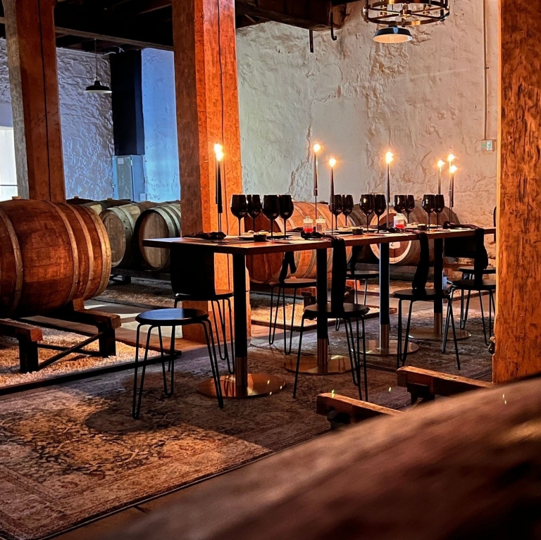 In a dim, atmospheric cellar at Hardys Tintara, McLaren Vale, South Australia, candlelit tables are set for a wine tasting. Large oak barrels line the historic stone walls, creating an intimate and rustic mood for this premium experience. © Hardys Tintara