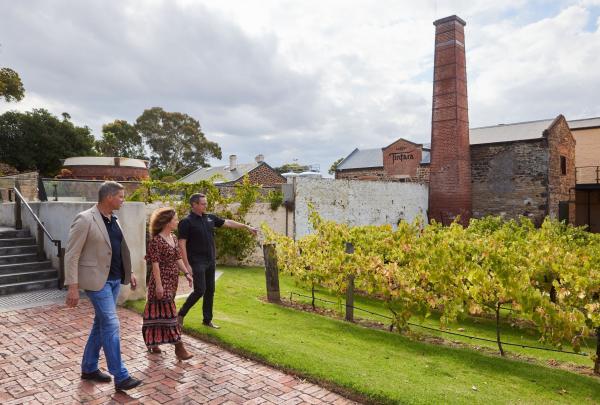 A guide leads a couple past a small vineyard at Hardys Tintara in McLaren Vale, South Australia. The historic stone winery buildings and a tall brick chimney stand under a cloudy sky, showcasing the heritage of this iconic wine region. © Hardys Tintara