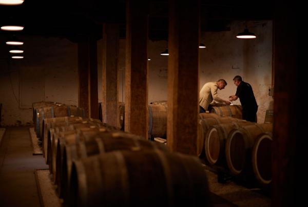 In a dimly lit cellar at Hardys Tintara in McLaren Vale, South Australia, two winemakers inspect large oak barrels. The warm, moody lighting highlights the historic timber beams and rows of maturing wine in this iconic winery. © Hardys Tintara