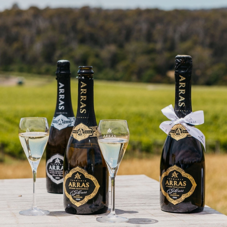 Three bottles of House of Arras sparkling wine and two filled glasses sit on a rustic wooden table. In the background, the rolling green vineyard of the Pipers River estate stretches toward the hills near Launceston, Tasmania. © House of Arras