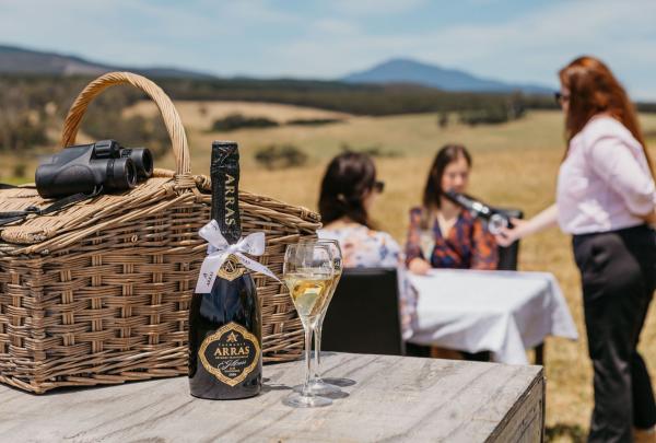 A bottle of House of Arras sparkling wine and two filled glasses sit on a rustic wooden table next to a picnic basket and binoculars. In the blurred background, guests enjoy a tasting overlooking the Pipers River hills near Launceston. © House of Arras