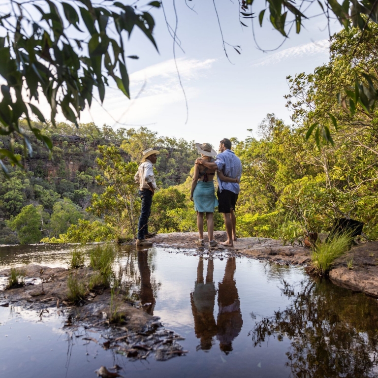 A guide from Jarramali Rock Art Tours stands with a couple of guests beside a beautiful waterfall, Cook, Queensland © Tourism Australia