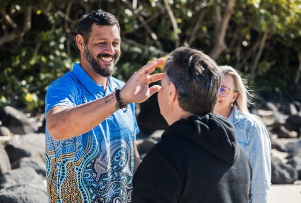 A friendly guide from the Jellurgal Aboriginal Cultural Centre applies traditional white face paint in intricate patterns to a smiling tour group participant, Walkabout Tour, Jellurgal Aboriginal Cultural Centre, Burleigh Heads, Queensland © Tourism Australia